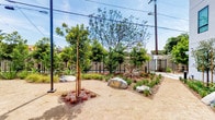 Located at 8740 S La Tijera Blvd in the Westchester neighborhood of Los Angeles, this single-story outdoor space is thoughtfully designed with a California-friendly landscape aesthetic. The grounds feature decomposed granite pathways that wind through carefully arranged planting beds filled with drought-tolerant vegetation, including ornamental grasses, agaves, and young shade trees. Natural boulders are strategically placed throughout the landscape, adding textural contrast and visual interest to the overall design.

The perimeter is defined by sleek, dark metal fencing that provides security while maintaining an open, airy feel. Accessible ramp entries with metal handrails ensure ease of movement throughout the space. Tall palm trees punctuate the landscape, lending a distinctly Southern California character, while additional trees offer growing canopy coverage.

The outdoor area sits adjacent to a modern multi-story building with a smooth gray stucco exterior and large windows, providing an urban backdrop to the naturalistic landscaping. Pathway lighting posts are positioned throughout the grounds, ensuring usability during evening hours. The overall ambiance is clean, contemporary, and low-maintenance, well-suited to the sunny Los Angeles climate.
