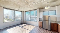 Located on the sixth floor at 707 Tenth Ave in the heart of downtown San Diego, this urban unit showcases a distinctive industrial-modern aesthetic. The open-plan living and kitchen area is defined by exposed concrete ceilings with visible ductwork, warm hardwood-style flooring, and expansive corner windows that flood the space with natural light and frame sweeping city skyline views. The kitchen is well-appointed with stainless steel appliances — including a refrigerator, range, microwave, and dishwasher — complemented by granite countertops, warm wood cabinetry on the lower level, and crisp white upper cabinets. A pendant light adds a finishing touch above the kitchen area. A hallway with louvered doors connects the main living space to the private rooms, including a bedroom featuring floor-to-ceiling windows with direct access to a balcony and an open closet with built-in rod. The bathroom is equipped with a corner glass-enclosed shower, granite vanity, and wood-toned cabinetry, consistent with the finishes throughout. Clean white walls and abundant natural light create a bright, open ambiance throughout this well-designed downtown San Diego residence.