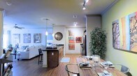 This single-floor space showcases an open-concept design that integrates the kitchen, dining, and living areas into one cohesive, flowing environment. The kitchen features a dark-topped island with seating, warm wood cabinetry, stainless steel appliances, and is illuminated by elegant pendant lights that draw the eye through the space. Track lighting along the ceiling provides additional brightness, complementing the warm hardwood-style flooring that runs throughout.

The dining area, positioned in the foreground, accommodates a rustic wood table with metal-accented chairs, adding an industrial touch to the overall aesthetic. A large indoor plant adds a natural, organic element alongside the decorative wall art that brings personality and color to the neutral-toned walls. Crown molding along the upper edges of the walls adds a refined architectural detail.

The living area is visible beyond the island, featuring a light-toned sofa arrangement in a bright, welcoming setting. The open layout maximizes the sense of space, creating an airy, connected atmosphere ideal for both everyday living and entertaining. Overall, the space balances modern design with warm, inviting accents.