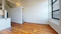 Located at 166 Valley St in Providence, Rhode Island, this two-floor loft-style space showcases a compelling blend of industrial heritage and contemporary updates. The main level greets visitors with rich, reclaimed hardwood floors that run throughout, crisp white walls, and soaring ceilings with exposed ductwork and structural elements that speak to the building's storied past. A pendant light anchors the open living area, which flows seamlessly into a well-appointed kitchen featuring granite countertops, light wood cabinetry, stainless steel appliances including a gas range, refrigerator, microwave, and dishwasher, as well as a modern pull-down faucet. A full bathroom on this level includes a tub-shower combination, tile flooring, and a granite-topped vanity. A staircase leads to the upper floor, where plush carpeting, a paneled ceiling with exposed beams, and large multi-pane windows invite natural light throughout. This level offers generous room for a bedroom suite, complete with dual closets providing ample storage. A second full bathroom on this floor mirrors the clean, functional design found below. The overall ambiance is that of a thoughtfully converted industrial loft, balancing original architectural details with modern comforts.