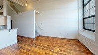 Located at 166 Valley St in Providence, Rhode Island, this two-floor loft-style space showcases a compelling blend of industrial heritage and contemporary updates. The main level greets visitors with rich, reclaimed hardwood floors that run throughout, crisp white walls, and soaring ceilings with exposed ductwork and structural elements that speak to the building's storied past. A pendant light anchors the open living area, which flows seamlessly into a well-appointed kitchen featuring granite countertops, light wood cabinetry, stainless steel appliances including a gas range, refrigerator, microwave, and dishwasher, as well as a modern pull-down faucet. A full bathroom on this level includes a tub-shower combination, tile flooring, and a granite-topped vanity. A staircase leads to the upper floor, where plush carpeting, a paneled ceiling with exposed beams, and large multi-pane windows invite natural light throughout. This level offers generous room for a bedroom suite, complete with dual closets providing ample storage. A second full bathroom on this floor mirrors the clean, functional design found below. The overall ambiance is that of a thoughtfully converted industrial loft, balancing original architectural details with modern comforts.