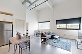Located at 3511 Tire Town Rd in Winston-Salem, NC, this single-story home offers a striking industrial-modern aesthetic across a well-planned layout. The open-concept kitchen and living room serves as the heart of the home, boasting dramatically high ceilings with exposed ductwork, polished concrete floors, and large glass-block and standard windows that flood the space with natural light and views of the surrounding landscape. The kitchen features white cabinetry, stainless steel appliances, and a breakfast bar, flowing seamlessly into the dining and living areas. A long, light-filled hallway connects the main living space to the private quarters, accented with wall sconces and art-ready white walls. The generously sized primary bedroom continues the clean, minimalist design with soaring ceilings and concrete flooring. A second bedroom offers a similarly spacious and bright atmosphere. The dedicated office provides ample room for a full workstation and lounge seating, with a glass-block window for natural light. The bathroom is crisp and modern, featuring a dark vanity with an illuminated mirror, a tub-shower combo with white subway tile surround, and polished fixtures. Additional spaces include four closets, a laundry area, and a hallway, completing this thoughtfully designed home.