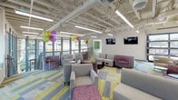 This resident lounge is a spacious, single-floor community gathering space designed to encourage socializing and relaxation. The open layout is anchored by a bold, contemporary aesthetic, featuring an exposed ceiling with industrial-style ductwork and linear pendant lighting that illuminates the entire floor. A striking combination of deep plum accent walls and bright, patterned carpet tiles in yellow, grey, and blue gives the space a lively, energetic character.

The main lounge area is generously furnished with clusters of comfortable grey and purple upholstered armchairs, loveseats, and small side tables, creating multiple intimate seating arrangements suited for both individual use and group gatherings. Large floor-to-ceiling windows along the perimeter bring in abundant natural light and offer open views of the surrounding outdoor greenery, enhancing the welcoming ambiance. Wall-mounted flat-screen televisions are positioned throughout for entertainment.

A compact but well-appointed kitchen area includes stainless steel appliances — a full-size refrigerator, electric range, and microwave — alongside grey cabinetry and a mosaic tile backsplash, making the space functional for residents. A restroom with a clean, modern design features deep plum walls, tile accents, and a wide sink counter with natural light provided by surrounding windows.