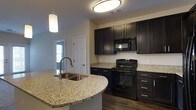 This single-floor space showcases a thoughtfully designed interior with a standout kitchen at its heart. The kitchen features rich, dark cabinetry that extends from floor to ceiling, providing ample storage while lending a bold, contemporary aesthetic. Granite countertops with a light speckled finish run throughout, offering both elegance and durability. A curved kitchen island takes center stage, fitted with a stainless steel sink and pendant lighting above, creating a natural gathering point that also serves as a functional workspace. Black appliances, including a range and over-the-range microwave, complement the dark cabinetry for a cohesive, modern look. Hardwood-style flooring anchors the space with warmth, while neutral-toned walls keep the atmosphere bright and open. A large overhead light fixture provides ample illumination in the kitchen, supplemented by natural light filtering in from a nearby window visible in the background. A hallway leading from the kitchen hints at additional rooms beyond, and a paneled interior door adds a classic architectural touch. The overall ambiance is clean, modern, and move-in ready, balancing style with everyday practicality.