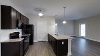 This single-floor space showcases an open-concept layout that seamlessly connects the kitchen and living area, creating a spacious and functional environment. The kitchen is a standout feature, centered around a large island with granite countertops and a double basin sink, illuminated by two sleek pendant lights that hang from the ceiling. Dark espresso-toned cabinetry lines the kitchen walls, providing ample storage, while black appliances — including a refrigerator, range, and overhead microwave — lend a cohesive, modern aesthetic. A flush-mount light fixture further brightens the cooking space. The flooring throughout is a warm hardwood-style material that adds continuity and visual warmth across the entire space. Neutral gray walls keep the atmosphere clean and versatile, complementing the darker tones of the cabinetry and appliances. The kitchen opens directly into the living area, which benefits from the natural flow of the layout. A white paneled interior door provides access to an additional room or utility space. Overall, the design balances functionality and contemporary style in a well-organized, single-level floor plan.