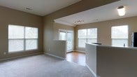 This single-floor space showcases an open-concept design that seamlessly connects the living area, dining space, and kitchen. The living room is finished with plush carpeting in a neutral tone, complemented by warm beige walls that create a welcoming ambiance throughout. A partial wall with a countertop bar separates the kitchen from the main living area, providing a functional and social transition between spaces. The kitchen bar features a clean, light-colored countertop surface ideal for casual dining or meal preparation.

The flooring transitions to hardwood in the dining and entryway area near the back door, adding a visual contrast and durability in high-traffic zones. Multiple windows, including a large window on the side wall and two additional windows flanking the back door, allow generous natural light to fill the space. The back door opens to what appears to be a covered balcony or patio, extending the living experience outdoors.

Ceiling details include a recessed section with a modern track lighting fixture, adding both depth and functional illumination. White trim and baseboards provide clean architectural definition throughout, enhancing the overall polished and well-maintained character of the space.