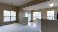 This single-floor space showcases an open-concept design that seamlessly connects the living area, dining space, and kitchen. The living room is finished with plush carpeting in a neutral tone, complemented by warm beige walls that create a welcoming ambiance throughout. A partial wall with a countertop bar separates the kitchen from the main living area, providing a functional and social transition between spaces. The kitchen bar features a clean, light-colored countertop surface ideal for casual dining or meal preparation.

The flooring transitions to hardwood in the dining and entryway area near the back door, adding a visual contrast and durability in high-traffic zones. Multiple windows, including a large window on the side wall and two additional windows flanking the back door, allow generous natural light to fill the space. The back door opens to what appears to be a covered balcony or patio, extending the living experience outdoors.

Ceiling details include a recessed section with a modern track lighting fixture, adding both depth and functional illumination. White trim and baseboards provide clean architectural definition throughout, enhancing the overall polished and well-maintained character of the space.