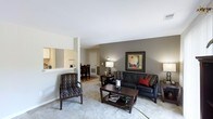 This single-floor space showcases an open-concept layout that seamlessly connects the living room, dining area, and kitchen. The living room serves as the focal point, featuring plush carpeted flooring, warm neutral wall tones, and a tasteful combination of a dark leather sofa accented with red throw pillows and patterned accent chairs. A wooden coffee table and coordinating side tables anchor the seating arrangement, while framed artwork and ambient table lamps add warmth to the space.

A half-wall partition separates the living area from the kitchen, providing a visual boundary while maintaining the open feel. The dining area, visible in the background, accommodates a dark wood dining set and benefits from natural light filtering through nearby windows. The kitchen features light cabinetry and is accessible via the pass-through counter along the partition wall.

The space is unified by consistent neutral carpeting throughout, smooth white ceilings, and a cohesive color palette of grays and warm taupes. The layout is practical and well-proportioned, offering a comfortable flow between functional areas suited to everyday living and casual entertaining.