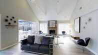 This single-floor space is designed to impress, combining functional and social areas under dramatically high ceilings with clean, modern finishes throughout. The entry lobby opens into a bright, expansive reception area featuring exposed white roof trusses and light hardwood flooring that runs seamlessly across the space. Recessed lighting and large French doors at the entrance flood the area with natural light, creating an airy, welcoming atmosphere. A dedicated workstation area is positioned off to one side, equipped with desks and office equipment, making the space well-suited for administrative or business center use.

Beyond the lobby, a sophisticated lounge area showcases soaring vaulted ceilings accented by track lighting. A central brick fireplace serves as the focal point of the room, flanked by large windows fitted with white blinds that allow generous natural light to filter in. Sliding glass doors connect the lounge to an outdoor patio, further enhancing the sense of openness. The lounge is furnished with a bold black leather sofa, plush accent chairs, and layered textiles, striking a balance between comfort and contemporary style. Light hardwood flooring and neutral wall tones tie both areas together, contributing to the space's cohesive and polished overall aesthetic.