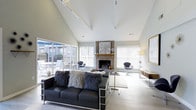 This single-floor space is designed to impress, combining functional and social areas under dramatically high ceilings with clean, modern finishes throughout. The entry lobby opens into a bright, expansive reception area featuring exposed white roof trusses and light hardwood flooring that runs seamlessly across the space. Recessed lighting and large French doors at the entrance flood the area with natural light, creating an airy, welcoming atmosphere. A dedicated workstation area is positioned off to one side, equipped with desks and office equipment, making the space well-suited for administrative or business center use.

Beyond the lobby, a sophisticated lounge area showcases soaring vaulted ceilings accented by track lighting. A central brick fireplace serves as the focal point of the room, flanked by large windows fitted with white blinds that allow generous natural light to filter in. Sliding glass doors connect the lounge to an outdoor patio, further enhancing the sense of openness. The lounge is furnished with a bold black leather sofa, plush accent chairs, and layered textiles, striking a balance between comfort and contemporary style. Light hardwood flooring and neutral wall tones tie both areas together, contributing to the space's cohesive and polished overall aesthetic.