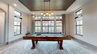 Located at 8050 Lapis Ln NW in Concord, NC, this single-floor space is thoughtfully designed for entertainment and leisure, combining modern transitional aesthetics with high-end architectural details throughout. The centerpiece of the space is a generously sized billiards room, anchored by a solid wood pool table set beneath a striking tray ceiling finished with dark shiplap wood paneling. A multi-pendant geometric chandelier adds a refined focal point, while floor-to-ceiling black-framed windows with transom panes flood the room with natural light and offer views of the surrounding tree-lined landscape. Soft, textured carpet in a cool marble-like tone grounds the room and flows seamlessly into the adjacent lounge area. The game lounge features high-top tables and bar-height seating, creating an inviting atmosphere for socializing, complemented by additional large windows and sliding glass doors that connect the interior to the outdoors. An entry corridor showcases intricate parquet hardwood flooring, arched niches with stone accent walls, and built-in cabinetry with marble countertops, adding a layer of architectural sophistication. Together, these spaces deliver a cohesive, upscale environment ideal for recreation and entertaining.