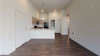 This single-floor residence is designed with a clean, contemporary aesthetic that emphasizes open living and functional design. The open-concept layout connects the kitchen and living area seamlessly, anchored by soaring ceilings that lend an airy, expansive feel to the space. Rich, dark hardwood-style flooring runs throughout the main living areas, providing a warm contrast to the bright white walls.

The kitchen is a standout feature, showcasing light wood cabinetry with sleek hardware, granite-look countertops, and a peninsula with an integrated sink — ideal for both meal prep and casual dining. Black appliances, including a range, microwave, and refrigerator, add a bold, modern accent. Pendant lighting above the peninsula completes the look.

Tucked conveniently near the kitchen is an in-unit laundry closet, housing a washer and dryer behind double doors for easy access without sacrificing living space.

The living area is bright and generously proportioned, featuring large windows that invite ample natural light. The bedroom offers plush carpeting underfoot, a window with natural light, and elevated ceilings that reinforce the sense of space.

The bathroom is finished with a full-size soaking tub and shower combination, chrome fixtures, and wood-style flooring that ties back to the rest of the home's cohesive design palette.