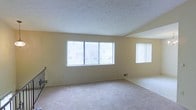 This single-floor space features an open living area designed to maximize natural light and comfort. The room is dressed in neutral, warm-toned walls that complement the soft, wall-to-wall carpeting underfoot, creating a welcoming and understated atmosphere throughout.

A prominent trio of windows along the main wall, fitted with vertical blinds, allows generous amounts of natural light to fill the space while offering views of the surrounding exterior. An additional side window further enhances the brightness and provides added ventilation options. Together, these windows give the room an airy, open feel.

The textured ceiling adds a subtle architectural detail that contrasts gently with the smooth walls below. A floor-mounted heating vent is positioned along the baseboard, indicating a built-in climate control system for year-round comfort. Partial visibility of a staircase railing on one side suggests access to additional areas or a split-level transition within the unit.

Overall, the space presents a clean, neutral canvas well-suited for a variety of furnishing styles, offering a functional and light-filled environment with practical features throughout.