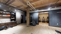 This single-floor fitness center showcases a striking combination of rustic charm and industrial character. The space is defined by its warm, exposed wood plank ceiling supported by heavy timber columns, which contrast beautifully with the exposed ductwork and utility piping overhead — a hallmark of industrial-inspired design. Light hardwood flooring runs throughout, enhancing the open, airy feel of the room.

The workout area is thoughtfully arranged to accommodate a variety of exercise needs. A multi-functional cable and resistance training machine is positioned centrally against a bold, dark patterned feature wall, creating a strong visual focal point. Adjacent to this, a row of cardio equipment — including treadmills equipped with personal display screens — lines the far end of the space, set against a contrasting neutral-toned wall.

Additional amenities include a built-in cabinetry unit with a countertop, ideal for storing supplies or providing a refreshment station. A private room, accessible via a dark-paneled door, offers additional functionality, potentially serving as a restroom or changing area. The overall ambiance is energetic yet refined, blending natural materials with purposeful fitness design to create an inviting and fully functional workout environment.