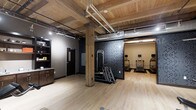 This single-floor fitness center showcases a striking combination of rustic charm and industrial character. The space is defined by its warm, exposed wood plank ceiling supported by heavy timber columns, which contrast beautifully with the exposed ductwork and utility piping overhead — a hallmark of industrial-inspired design. Light hardwood flooring runs throughout, enhancing the open, airy feel of the room.

The workout area is thoughtfully arranged to accommodate a variety of exercise needs. A multi-functional cable and resistance training machine is positioned centrally against a bold, dark patterned feature wall, creating a strong visual focal point. Adjacent to this, a row of cardio equipment — including treadmills equipped with personal display screens — lines the far end of the space, set against a contrasting neutral-toned wall.

Additional amenities include a built-in cabinetry unit with a countertop, ideal for storing supplies or providing a refreshment station. A private room, accessible via a dark-paneled door, offers additional functionality, potentially serving as a restroom or changing area. The overall ambiance is energetic yet refined, blending natural materials with purposeful fitness design to create an inviting and fully functional workout environment.
