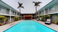 This property centers around a generous outdoor courtyard pool area that serves as the focal point of the complex. The pool itself is expansive, with clear turquoise water and a wide surrounding deck that provides ample space for lounging. Tall palm trees rise prominently at the corners of the courtyard, lending a distinctly tropical, resort-like atmosphere to the space.

The courtyard is framed on multiple sides by a two-story building with an L- or U-shaped layout, featuring covered walkway balconies on the upper level that overlook the pool area. The exterior architecture has a clean, mid-century influenced design with neutral tones and consistent railings throughout.

Poolside amenities include a covered cabana with warm-toned draping, as well as multiple lounge chairs and patio seating arrangements shaded by large market umbrellas. Tropical landscaping, including low shrubs and ornamental plants, lines the perimeter, adding greenery and visual depth to the courtyard. The overall ambiance is relaxed and inviting, evoking a Southern California coastal aesthetic suited for leisure and outdoor enjoyment.
