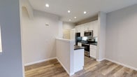 This single-floor space showcases a clean, modern design with an open and airy layout that promotes seamless flow between living areas. The kitchen serves as a central highlight, featuring bright white shaker-style cabinetry that extends to the upper walls, providing ample storage throughout. Dark countertops offer a striking contrast against the white cabinetry, while a peninsula counter extends outward to define the kitchen space and create an informal eating or prep area. Stainless steel appliances — including a refrigerator, microwave, and range — complete the contemporary kitchen aesthetic. Recessed lighting is installed throughout the ceiling, ensuring consistent, well-distributed illumination across the space. The flooring is a warm hardwood-style material that runs throughout, adding texture and warmth to the otherwise neutral palette of light gray walls and crisp white trim. A louvered closet door is visible in the background, hinting at practical storage solutions integrated into the design. Overall, the space presents a well-appointed, move-in-ready interior with a cohesive modern style, combining functionality with clean architectural lines and a neutral color scheme that offers versatility for a variety of furnishing choices.