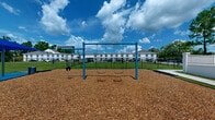 Located in Baton Rouge, Louisiana, this outdoor playground offers a thoughtfully designed recreational space for children of all ages. The play area is surfaced throughout with a generous layer of wood chip mulch, providing a safe and impact-absorbing ground cover beneath all equipment.

A prominent blue swing set serves as a centerpiece of the space, featuring multiple standard belt swings alongside a bucket-style toddler swing, accommodating a range of ages simultaneously. Nearby, an eye-catching interactive play structure shaped like a friendly character adds imaginative play value to the environment.

A blue shade canopy covers an adjacent seating area equipped with a picnic table, offering a comfortable, shaded spot for caregivers and visitors. Additional ground-level interactive play panels and activity stations are positioned throughout, encouraging sensory and developmental engagement.

The playground is enclosed by a sturdy black metal fence with white masonry columns, creating a secure perimeter while maintaining open sightlines to the surrounding landscape. Mature pine trees and lush green lawn frame the space, and views of neighboring buildings and a commercial high-rise beyond the treeline provide an urban-suburban backdrop.

The open setting benefits from abundant natural light under the characteristic Louisiana sky, making it an inviting and functional outdoor destination for the surrounding community.