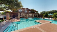 Located at 8690 Virginia Pkwy in McKinney, Texas, this two-floor property features a striking resort-style outdoor amenity area centered around a generously sized swimming pool. The complex showcases classic Texas architecture with warm brick and stone exteriors, arched windows, and a distinctive cupola-topped clubhouse building clad in floor-to-ceiling glass panels, allowing abundant natural light to fill the interior.

The pool area is a standout feature, designed for both relaxation and recreation. A decorative pergola structure with stone columns serves as a dramatic focal point, complete with cascading water features that add both visual appeal and ambient sound. The pool deck is finished in rich terracotta-toned concrete and is furnished with lounge chairs, umbrellas, and casual seating areas, offering ample space for sunbathing and outdoor dining.

Mature shade trees are thoughtfully integrated throughout the grounds, providing natural canopy and a lush, park-like atmosphere. The perimeter is enclosed by wrought iron fencing, ensuring privacy and security. The two-story residential buildings flanking the pool area feature warm brick facades, pitched rooflines, and private balconies, contributing to the cohesive, upscale aesthetic of the overall community.