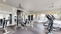 Located in Largo, Florida, this fitness center offers a thoughtfully designed exercise space that balances functionality with an inviting atmosphere. The room features neutral-toned walls accented by crisp white crown molding and trim, lending a polished, upscale feel to the workout environment. Large windows line multiple walls, flooding the space with abundant natural light and offering pleasant views of the surrounding lush greenery and outdoor amenity areas, including glimpses of a pool. Dual ceiling fans ensure consistent air circulation throughout the room, enhancing comfort during workouts. The durable, cushioned flooring provides a practical and supportive surface for a wide variety of exercises. Equipment is thoughtfully arranged to maximize use of the available floor space and includes treadmills, elliptical trainers, a recumbent bike, rowing machines, cable weight machines, a multi-station strength system, and a dumbbell rack. Full-length mirrors are positioned along one wall, amplifying the sense of space and allowing users to monitor their form. Exercise mats and free weights round out the offerings. The overall ambiance is clean, bright, and well-maintained, making this fitness center a functional and appealing facility for daily exercise.