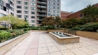 Located at 4400 East-West Hwy in Bethesda, Maryland, this outdoor courtyard offers a well-designed open-air retreat nestled within an urban setting. The space is defined by a broad, light-toned tiled terrace that provides ample room for movement and relaxation. At its centerpiece sits a raised brick fountain basin with multiple water jets, adding both visual interest and a calming auditory element to the environment. Surrounding the terrace are generously proportioned raised planter beds constructed from light brick, filled with a rich variety of mature trees, shrubs, ornamental grasses, and seasonal plantings that provide lush greenery and natural enclosure. Strategically placed metal benches in a dark green finish offer comfortable seating spots throughout the courtyard. The landscaping includes a layered design, with taller trees providing a natural canopy and lower-growing plants filling the planters with color and texture. The courtyard is framed by the façade of a multi-story residential high-rise, whose balconies overlook the space, as well as adjacent brick commercial buildings. The overall ambiance is tranquil and well-maintained, making it a welcoming outdoor space suited for quiet rest or casual gathering in the heart of Bethesda.