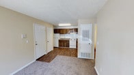 This single-floor residence offers a straightforward and functional layout designed for comfortable everyday living. The living area is finished with wall-to-wall carpet, providing a warm and comfortable underfoot feel, and flows seamlessly into the kitchen area, which transitions to durable hardwood-style flooring. The kitchen is equipped with dark wood cabinetry offering ample storage, a white electric range, a refrigerator, and a sink positioned beneath the upper cabinets. A fluorescent light fixture provides bright, even illumination over the kitchen workspace. The neutral, light-toned walls throughout the space lend a clean and airy feel, complemented by a textured popcorn ceiling that adds a classic touch. Two white panel doors are visible near the entry area, likely leading to a closet and an exterior or utility access point, while an additional door on the opposite side suggests access to further rooms or a private exterior entry. A floor-level ventilation unit adds to the practical, no-fuss character of the space. Overall, this unit presents a tidy, functional environment well-suited for those seeking a simple and manageable living space.