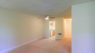 This single-floor space offers a straightforward, functional layout designed for comfortable everyday living. The main living area is generously sized, finished with wall-to-wall beige carpeting that adds warmth underfoot. A ceiling fan with an integrated light fixture is centrally mounted on the textured popcorn ceiling, providing both air circulation and ambient lighting. Neutral-toned walls throughout create a clean, versatile backdrop.

A wall opening connects the living area directly to the kitchen, which features light wood cabinetry, a white dishwasher, and a sink positioned beneath upper cabinets. The kitchen is finished with smooth flooring, contrasting with the carpeted living spaces. A wall-mounted HVAC vent near the base of the wall ensures climate control in the main living area.

A hallway visible from the living space suggests additional rooms beyond, hinting at a thoughtful floor plan with clear separation between living and private areas. The overall ambiance is clean and neutral, offering a move-in-ready environment with classic, understated finishes throughout.
