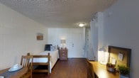 This single-floor studio room offers a practical and straightforward layout designed to maximize functionality within a compact footprint. The space features dark hardwood-style flooring that contrasts with the white-painted concrete block walls, creating a clean and utilitarian aesthetic throughout.

The sleeping area is furnished with a twin-sized loft-style wooden bed frame, accompanied by a wooden nightstand with drawers for convenient bedside storage. A small desk lamp on the nightstand provides soft ambient lighting to complement the ceiling-mounted flush light fixture overhead.

On the opposite side of the room, a built-in open closet area is partially concealed by a white curtain on a grommet rod, offering storage for clothing and personal items. Shelving within the closet provides additional organizational options.

A wooden study desk positioned near the closet area is equipped with a task lamp, making it a dedicated workspace for reading or studying. The room's textured popcorn ceiling and white-toned walls contribute to a neutral, no-frills atmosphere. A small piece of wall art adds a modest decorative touch. The entrance door is centrally located along the back wall, providing straightforward access to the space.