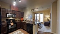 This single-floor space showcases an open-concept layout that integrates the kitchen and living area into one cohesive, functional environment. The kitchen is a standout feature, equipped with rich dark wood cabinetry, dark countertops, and black appliances, including a range and built-in microwave. A peninsula with a sink and an eat-in overhang adds both workspace and casual dining potential, serving as a natural divider between the kitchen and living area. Track lighting runs across the ceiling, providing ample illumination throughout the space. The flooring transitions from hardwood-style planks in the kitchen to a carpeted living area, adding textural contrast. The living area benefits from natural light streaming through double windows and a glass-paneled door, creating a bright and welcoming atmosphere. Neutral beige walls complement the warm tones of the cabinetry and furnishings. A decorative accent wall near the kitchen adds visual interest with its earthy tone. Overall, the space offers a comfortable and practical layout with a warm, inviting ambiance suited for everyday living.