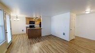 This single-floor space is designed with an open-concept layout that seamlessly connects the kitchen and living area, creating a functional and cohesive environment. Light hardwood-style flooring runs throughout the main living space, contributing to a clean and contemporary aesthetic.

The kitchen is positioned toward the rear of the open area and is defined by warm honey-toned wood cabinetry that provides ample storage. A peninsula with a dark wood-paneled base and granite-style countertop separates the kitchen from the living space, offering additional prep or seating space. Black appliances, including a range and microwave, are integrated into the cabinetry, and track lighting overhead ensures the kitchen is well-lit.

The living area is spacious and open, with white walls that create a bright, neutral backdrop. The smooth white ceiling enhances the sense of openness throughout. A wall-mounted thermostat and utility access panel are visible on one wall, and a paneled door provides access to an adjacent room or exterior. The overall ambiance is clean, practical, and move-in ready.
