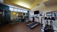 This fitness center is thoughtfully designed to accommodate a full range of exercise needs within a single, open-plan space. The room features vaulted ceilings that lend an airy, spacious quality, while a band of large windows along one wall allows natural light to fill the interior and offers views of the surrounding outdoor landscape.

The cardio section is well-stocked with multiple True-brand treadmills, an elliptical trainer, and a stair climber, providing a variety of aerobic workout options. A wall-mounted flat-screen television is positioned for easy viewing during cardio sessions. For strength training, the facility includes a cable weight machine and an adjustable workout bench, along with a dumbbell rack offering a range of free weights. A water cooler is conveniently available for hydration.

A large mirror spans one wall, visually expanding the space and allowing users to monitor their form during exercise. The floor is finished with durable, dark carpet suited for fitness use. Informational boards and workout charts are posted on the walls, adding a functional, resource-oriented element to the environment. The overall ambiance is practical and well-maintained, supporting an effective fitness experience.