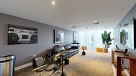 Located on Biscayne Boulevard in North Miami Beach, this music-inspired lounge room offers a unique and well-appointed space designed for both relaxation and creative expression. The room is finished with textured gray walls, recessed lighting, and wall-to-wall carpeting that lends a warm, acoustic-friendly atmosphere throughout.

At the heart of the space is a full electronic drum kit and a keyboard on a stand with a bench, making it a functional environment for musicians. A sleek black leather sofa anchors the seating area, complemented by a round side table and vibrant yellow ottomans that add a bold pop of color. Music-themed artwork — including a striking black-and-white boombox print and a framed photograph of a performer — adorns the walls, reinforcing the room's creative identity.

A large flat-screen television is mounted on one wall above a wooden console, ideal for media viewing or entertainment. Floor-to-ceiling sheer white curtains line the exterior wall, diffusing natural light beautifully and offering a soft view of tropical greenery beyond. A tall indoor plant further enhances the fresh, lively ambiance of the space.

Practical elements include a spacious closet with louvered doors and a glass-paneled entry door. The overall design strikes a balance between stylish lounge comfort and a fully equipped creative studio.