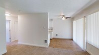 This single-floor space offers a clean, functional layout designed for comfortable everyday living. The main living area is generously sized, featuring a combination of plush carpeting and hardwood-style flooring that transitions naturally between zones. A ceiling fan with an integrated light fixture is centrally positioned overhead, providing both air circulation and ambient lighting. The textured ceiling adds a classic finish throughout the space.

A partial dividing wall separates the living area from the rest of the unit, creating a sense of definition without fully enclosing the floor plan. This open layout allows natural light to flow freely through the space, which is enhanced by vertical blinds covering what appears to be a sliding door or large window. The bright, neutral white walls create a clean and versatile backdrop throughout.

The kitchen, visible from the living area, features white cabinetry and appears compact yet functional. A wall-mounted heater vent in the living area ensures efficient climate control. A small pet door is also integrated into the wall, adding a practical touch for pet owners. The overall ambiance is light, airy, and move-in ready, making it a welcoming and adaptable space.