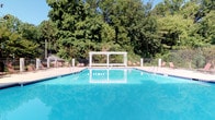 This outdoor recreational space centers around a generously sized swimming pool with crystal-clear blue water, designed to accommodate both leisurely and active use. A distinctive white pergola structure rises from the pool's center, serving as a visual focal point and functional accent to the overall layout. The pool is equipped with multiple entry points via stainless steel handrails, ensuring safe and convenient access.

Surrounding the pool, a spacious concrete deck provides ample room for relaxation, lined with red lounge chairs positioned to take advantage of the open-air environment. The entire area is enclosed by a black metal safety fence, offering both security and a clean aesthetic boundary.

A small covered shelter or shade structure is visible on the far left of the deck, providing an additional retreat from direct sunlight. The backdrop of the space is defined by a dense canopy of mature trees, creating a natural, private, and serene atmosphere. Under clear blue skies, the setting feels open and inviting throughout the day. The combination of lush greenery, clean pool water, and well-arranged amenities makes this outdoor area a comfortable and functional recreational destination.