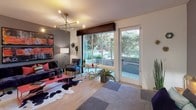 Located at 1980 Kettner in San Diego's vibrant Little Italy neighborhood, this model residence offers a thoughtfully designed single-floor layout that blends contemporary style with everyday functionality. The open-concept living area serves as the heart of the space, featuring warm hardwood-style flooring, a striking modern chandelier with brass accents, and a bold accent wall in deep charcoal grey. Large sliding glass doors and a full-length side door flood the room with natural light and provide direct access to an outdoor patio area surrounded by lush greenery, creating a seamless indoor-outdoor connection. The kitchen is a standout feature, boasting sleek dark grey cabinetry, a generous granite island with a waterfall edge and undermount sink, stainless steel appliances including a gas range, microwave, refrigerator, and dishwasher, and elegant pendant lighting overhead. Light hardwood-style flooring runs throughout, unifying the space. The bathroom continues the modern aesthetic with a charcoal accent wall, tile flooring, and a stylish framed mirror. Overall, the residence presents a cohesive, design-forward ambiance that balances bold color choices with clean, contemporary finishes throughout.