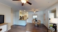 This single-floor residence showcases an open-concept floor plan that integrates the kitchen, dining, and living areas into one cohesive, flowing space. The interior is finished with rich, dark hardwood-style flooring that runs throughout, providing a warm contrast to the crisp white walls and ceilings. Ceiling fans are installed in both the living and dining zones, promoting airflow and comfort throughout the space.

The kitchen features white cabinetry, recessed lighting, and a peninsula with a dark countertop that provides additional workspace and casual seating. Stainless steel appliances, including a refrigerator and microwave, complete the functional cooking area. The peninsula acts as a natural divider between the kitchen and the adjacent living space without disrupting the open feel.

The dining area is positioned near a window that invites natural light into the room, creating a bright and welcoming atmosphere. Crown molding along the upper walls adds a refined architectural detail throughout the space. Overall, the layout offers a clean, functional, and comfortable living environment with a neutral palette suited to a variety of interior styles.