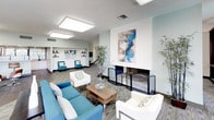 This single-story community clubhouse is thoughtfully designed to serve as a social and relaxation hub. At its heart is a spacious lounge area featuring soft, teal-toned sectional seating, accent armchairs, and a warm fireplace framed by contemporary artwork, all illuminated by large recessed light panels overhead. The color palette of soft greens, whites, and blues lends a calm, cohesive ambiance throughout.

An adjacent seating area opens to the outdoors through expansive floor-to-ceiling glass doors and windows, allowing abundant natural light to flood the interior while offering direct views of the surrounding landscaping and courtyard. This area transitions seamlessly into a bar counter lined with stylish swivel stools, a wall-mounted TV, and additional casual seating — ideal for gatherings or everyday use.

A well-equipped catering kitchen features dual sinks, generous counter space, under-counter appliances, and dark hardwood-style flooring, providing practical amenities for events.

Outside, the pool area is a standout feature, offering a shimmering swimming pool, a spa, shaded lounge seating under teal umbrellas, and lush palm trees — evoking a true resort atmosphere.