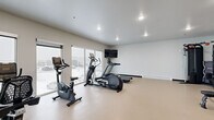 Located at 3911 20th Ave S in Fargo, North Dakota, this fitness center offers a well-equipped, single-room exercise space designed to support a well-rounded workout routine. The room features durable, light-toned flooring throughout and is illuminated by recessed ceiling lights, creating a bright and functional environment.

Large sliding windows line one wall, flooding the space with natural light and providing views of the surrounding area. A wall-mounted flat-screen television is positioned for easy viewing during workouts. The cardio equipment selection includes treadmills, elliptical trainers, and stationary bikes, all arranged to allow comfortable use of each machine.

For strength training, the facility includes a multi-station cable machine, a weight bench, and a dumbbell rack stocked with a range of free weights. A full-length wall mirror enhances the sense of space and allows users to monitor their form during exercise. A wall-mounted split air conditioning unit provides climate control year-round.

Near the entrance, a black-framed glass partition wall and door provide visibility into an adjacent space, while a dual-basin water fountain is conveniently available just outside the main exercise room. A medicine ball rack rounds out the amenities. The overall design is clean, modern, and purposefully laid out to maximize usability within the space.