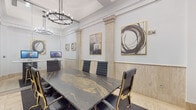 Located at 2121 S Hiawassee Rd in Orlando, Florida, this single-floor commercial space blends classical architectural elegance with modern functionality. The interior is defined by polished travertine marble flooring that extends throughout, complemented by an ornate mosaic tile inlay running along the main corridor — a striking design element that anchors the flow of the space.

The central hallway connects the key functional areas and is lined with full-height glass partition walls, offering transparency and openness while maintaining defined zones. Circular chandelier lighting fixtures suspended from coffered ceilings lend a refined, upscale ambiance across the entire space.

A formal conference room serves as a standout feature, appointed with a dramatic black-and-gold marble conference table surrounded by sleek black executive chairs with gold-accented frames. Tall decorative pilasters with stone wainscoting line the walls, and gold-framed abstract artwork adds a contemporary artistic touch. A wall-mounted monitor supports presentations and meetings.

Adjacent to the conference room, a glass-enclosed lounge or media area provides a more relaxed setting, equipped with a large flat-screen display, ambient blue accent lighting, and comfortable seating — ideal for client entertainment or informal gatherings. Together, these spaces create a professional yet distinctly luxurious environment suited for executive offices or client-facing operations.