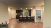 This single-floor space is designed around an open-concept layout that seamlessly integrates the kitchen and living areas. The kitchen serves as the focal point, showcasing dark espresso cabinetry that contrasts cleanly against the neutral beige walls. Stainless steel appliances, including a full-sized refrigerator, microwave, and range, lend a modern, polished aesthetic. A tall pantry cabinet provides additional storage, enhancing the kitchen's functionality. The centerpiece of the kitchen is a generously sized dark island with a sleek countertop, accommodating four cushioned bar stools—ideal for casual dining or entertaining. Recessed lighting is distributed evenly throughout the ceiling, ensuring consistent illumination across the space. The flooring features warm-toned hardwood-style planks that run throughout, adding visual continuity and a sense of warmth. A doorway to the right hints at an adjacent room, partially visible and offering a glimpse of natural light filtering through window blinds. The overall ambiance is contemporary and functional, with a cohesive dark-and-neutral color palette.