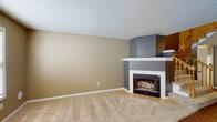 This two-floor residence offers a thoughtfully laid-out interior with a blend of practical and comfortable design elements throughout. The living area serves as a welcoming focal point, anchored by a corner fireplace framed in white trim and gray surround, with plush carpeting underfoot and a textured ceiling overhead. The kitchen is well-equipped, featuring white shaker-style cabinetry extending to the ceiling, granite-look countertops, hardwood-style flooring, and black appliances including a range, microwave, dishwasher, and refrigerator. A window above the sink allows natural light to filter into the space. The bedrooms are generously proportioned, each finished with soft carpeting, neutral warm-toned walls, ceiling fans with light fixtures, and windows fitted with horizontal blinds for privacy and light control. The bathrooms are cleanly finished, with granite-topped white vanities, tile flooring, and large mirrors. One bathroom includes accessibility features such as chrome grab bars, while another offers a full tub and shower combination. A staircase connects the two levels, contributing to the home's multi-story layout. Throughout the space, a consistent warm neutral color palette and clean white trim create a cohesive and inviting atmosphere.
