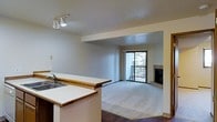 This single-floor space showcases an open-concept design that seamlessly connects the kitchen and living areas. The kitchen features a raised bar counter with warm wood trim edging and white laminate surfaces, complemented by a sink and track lighting overhead that illuminates the workspace effectively. The flooring transitions from dark hardwood in the kitchen area to plush carpeting in the living space, creating a natural visual boundary between the two zones.

The living area is generously proportioned, offering a comfortable and versatile layout. A standout feature is the tile-surround fireplace positioned along one wall, providing both a functional heating element and a focal point for the room. Sliding glass doors flood the living space with natural light and open onto a private balcony, offering views of surrounding greenery. The neutral, light gray wall tones throughout create a clean and cohesive aesthetic. A textured ceiling adds subtle dimension, while the open pass-through design between the kitchen and living room enhances the sense of spaciousness, making the overall layout feel connected and inviting.