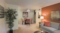 This single-floor residence is designed with an open, connected layout that allows the living and dining areas to flow seamlessly into one another. The dining space is anchored by a round glass-top table surrounded by upholstered chairs, set beneath a ceiling fan with integrated lighting — a practical and stylish centerpiece. A framed piece of wall art adds a decorative focal point to this area, while a mirrored closet in the background reflects light and enhances the sense of openness.

The living area features a bold terracotta accent wall that introduces warmth and visual depth to the space. A coordinating table lamp and side table provide functional ambient lighting, complementing the overall warm color palette. Plush wall-to-wall carpeting runs throughout, contributing to a comfortable, inviting atmosphere underfoot.

The textured ceiling adds a classic residential character, and the neutral beige tones on the primary walls provide a versatile backdrop that suits a variety of furnishing styles. Ample wall space and well-placed lighting make this a practical and welcoming home, suited for both everyday living and casual entertaining. The cohesive design choices throughout create a unified and comfortable single-level living environment.
