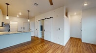 This single-floor space showcases a thoughtfully designed open layout with warm hardwood-style flooring that flows consistently throughout. The living area is spacious and bright, featuring white walls, ceiling fans, and recessed lighting that contribute to a clean, airy ambiance.

The kitchen is a standout feature, outfitted with a full suite of stainless steel appliances, including a refrigerator, range, microwave, and dishwasher. Tall white cabinetry extends nearly to the ceiling, maximizing storage, while granite countertops and a white subway tile backsplash add polished detail. A kitchen island with pendant lighting above provides additional workspace and a natural gathering point.

A distinctive black-hardware sliding barn door serves as a functional and stylish element, offering access to what appears to be a bathroom or utility area. The bathroom is clean and modern, featuring a white vanity with a simple countertop, continued hardwood-style flooring, and crisp white walls.

Additional features include a dedicated utility closet housing a water heater, louvered closet doors, and a separate hallway corridor providing clear spatial separation between areas. The overall design is contemporary and cohesive, with consistent finishes throughout.
