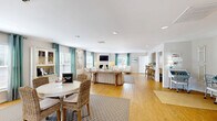 Located at 4632 Still Meadow Dr in Wilmington, NC, this single-floor community space offers a thoughtfully designed and inviting coastal atmosphere throughout. The open-concept living room serves as the heart of the space, showcasing warm hardwood flooring, recessed lighting, and an abundance of natural light streaming through multiple windows dressed in teal gypsy-curtains with plantation shutters. A classic white fireplace anchors the seating area, which is complemented by neutral toned sofas, tufted accent chairs, and a glass-top coffee table. A dining nook with a round white table and woven rattan chairs adds a relaxed, beachy charm, while a built-in display hutch provides functional storage. Coastal artwork and starfish wall accents reinforce the seaside aesthetic throughout. The hallway serves as a clean, well-lit transitional space connecting the main areas, featuring hardwood floors, recessed lighting, and glass-paned interior doors. The dedicated laundry room is fully equipped with multiple stacked commercial-grade washer and dryer units, a folding table, and wood-look flooring, offering a practical and organized shared facility. Together, these spaces create a comfortable, well-maintained community environment with a consistent coastal design theme.