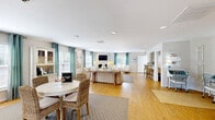 Located at 4632 Still Meadow Dr in Wilmington, NC, this single-floor community space offers a thoughtfully designed and inviting coastal atmosphere throughout. The open-concept living room serves as the heart of the space, showcasing warm hardwood flooring, recessed lighting, and an abundance of natural light streaming through multiple windows dressed in teal gypsy-curtains with plantation shutters. A classic white fireplace anchors the seating area, which is complemented by neutral toned sofas, tufted accent chairs, and a glass-top coffee table. A dining nook with a round white table and woven rattan chairs adds a relaxed, beachy charm, while a built-in display hutch provides functional storage. Coastal artwork and starfish wall accents reinforce the seaside aesthetic throughout. The hallway serves as a clean, well-lit transitional space connecting the main areas, featuring hardwood floors, recessed lighting, and glass-paned interior doors. The dedicated laundry room is fully equipped with multiple stacked commercial-grade washer and dryer units, a folding table, and wood-look flooring, offering a practical and organized shared facility. Together, these spaces create a comfortable, well-maintained community environment with a consistent coastal design theme.
