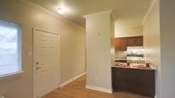 This single-story space is designed with a clean, functional layout that seamlessly connects the entryway to the kitchen area. The entryway is highlighted by warm beige walls, hardwood-style flooring, and a classic paneled closet door, providing both practicality and a welcoming atmosphere. Crown molding runs along the ceiling throughout the space, adding a refined architectural detail, while a textured ceiling and flush-mount light fixture complete the overhead design.

A distinctive structural column with a decorative cap separates the entryway from the kitchen, serving as both an architectural feature and a natural room divider. The kitchen showcases rich dark wood cabinetry paired with granite-style countertops and a mosaic tile backsplash, adding texture and visual interest. A peninsula counter with a dark base provides additional workspace and casual seating potential. White appliances and under-cabinet lighting contribute to the kitchen's bright, practical design. Overall, the space presents a cohesive aesthetic defined by warm tones, classic detailing, and thoughtful design elements.