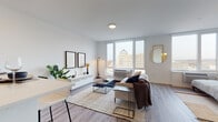 Located at 150 Main St in Hackensack, NJ, this modern residence offers a thoughtfully designed single-floor layout that blends contemporary aesthetics with functional living. The open-concept living area is anchored by large, floor-to-ceiling windows that flood the space with natural light and frame sweeping views of the Hackensack skyline, including the iconic domed building prominent in the cityscape. Light wood-toned flooring flows throughout, complementing the clean, neutral color palette.

The kitchen is outfitted with dark charcoal cabinetry, stainless steel appliances including a range, over-the-range microwave, and refrigerator, as well as crisp white quartz countertops and a classic white subway tile backsplash. A peninsula with bar seating provides additional dining and prep space, making it ideal for entertaining.

The sleeping area, visible in the dollhouse view, is comfortably proportioned and positioned adjacent to in-unit laundry facilities. The bathroom features marble-style tile flooring, a dark-toned vanity with a white countertop, a tiled shower-tub combination, and clean, minimalist fixtures. Throughout the residence, the design emphasizes a bright, airy ambiance with modern detailing that suits contemporary urban living.