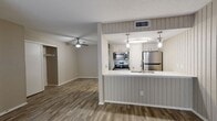 This contemporary apartment boasts a thoughtfully designed open floor plan. The kitchen showcases stainless steel appliances, white cabinetry, and pendant lighting. The living area is spacious and well-lit, with a large window offering outdoor views. Wood-style flooring extends throughout, complementing the neutral wall colors. The bathroom features a single vanity and a tub/shower combination. A ceiling fan in the bedroom adds comfort. Vertical paneling on some walls adds texture and visual interest. The overall design emphasizes functionality and a clean, modern aesthetic.