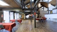This single-floor space blends industrial and rustic aesthetics to create a versatile gathering environment. Soaring ceilings with exposed steel beams and corrugated metal panels define the architectural character, lending the space a bold, open feel. Sturdy structural steel columns rise throughout the floor plan, reinforcing the industrial design while framing distinct zones within the open layout.

The hardwood flooring adds warmth and continuity across the space, contrasting beautifully with the raw metal and brick elements. A prominent brick feature wall on one side introduces texture and a sense of heritage, complementing the modern industrial framework above.

Natural light filters in through large windows along the perimeter, brightening the interior and offering views of the surrounding outdoor environment. A decorative pendant light fixture adds an elegant touch amid the rugged structural elements overhead.

The space is arranged with tables and chairs dressed in red linens, making it well-suited for events, banquets, or community gatherings. The open floor plan allows for flexible furniture configurations, accommodating a variety of functions and group sizes with ease.
